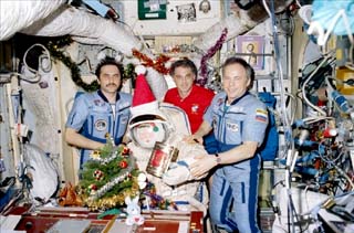 Mir-24 crewmembers posing with a santa doll in a Sokol suit, small tree, decked boughs, and floating presents.